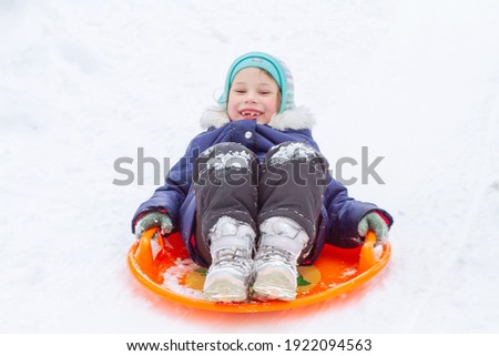 A little pretty girl in a overalls slides down an ice slide, winter children's games, a child in the park in winter.
