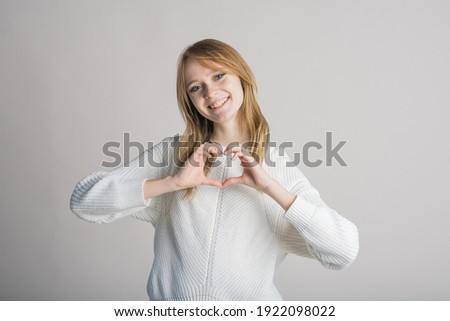 Attractive Caucasian young blonde woman in casual clothing looks at camera, smiles and shows gesture heart shape isolated on gray studio background. Love, feeling, romance, friendship.