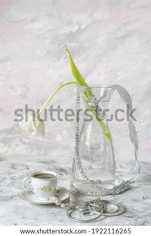 Delicate white tulip flower in a glass vase, a cup of coffee and various silver decorations on a light background. light still life.