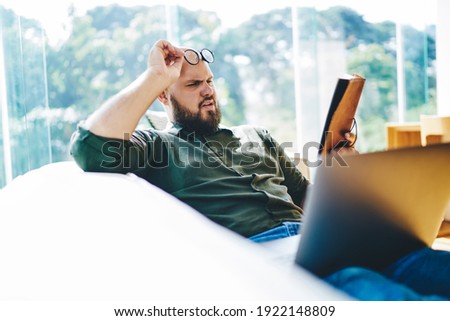 Side view of young man with beard reading book holding eyeglasses and squinting eyes for making better eyesight while resting on sofa with laptop