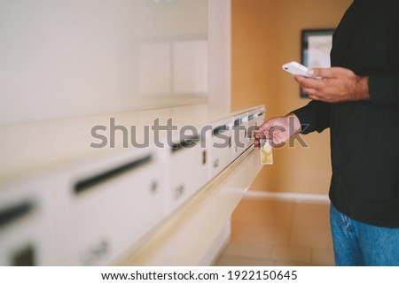 Side view of man in casual wear reading message on mobile phone while unlocking with key letter box in lounge of apartment building