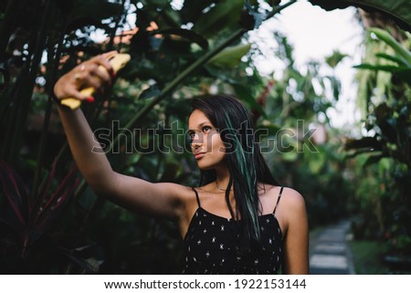 Positive young woman in stylish dress standing between green plants and taking selfie on mobile phone in tropical garden on daytime
