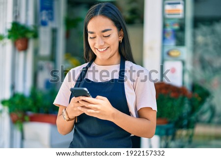 Young latin shopkeeper girl smiling happy using smartphone at florist. Royalty-Free Stock Photo #1922157332