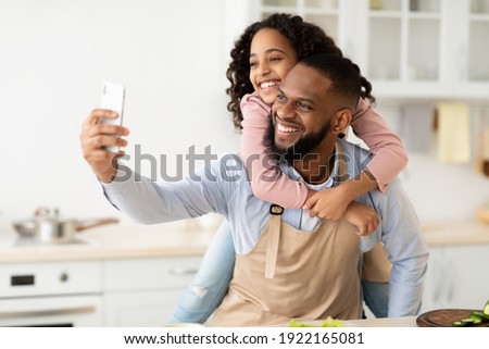 Fatherhood Concept. Portrait of cheerful african american father taking selfie with his little daughter while cooking in the kitchen together at home, giving her piggyback ride, using smartphone