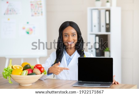 Positive female nutrition specialist pointing at laptop with blank screen at medical office, mockup for your website. Professional black dietitian promoting weight loss clinic, space for design