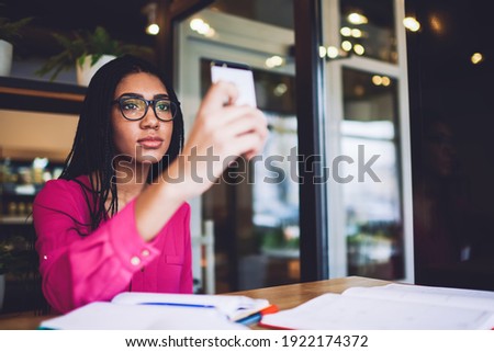 Concentrated African American lady with braids in casual wear and eyeglasses taking photo on smartphone while working with notes in cafe