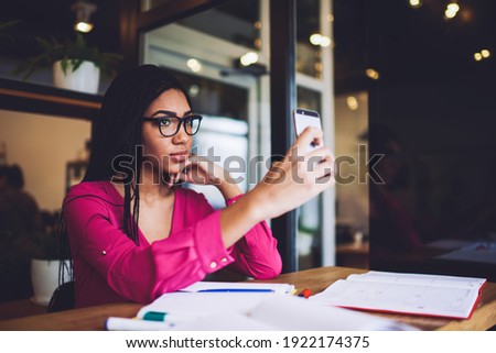 Elegant African American lady with braids in casual wear and spectacles taking selfie on smartphone while working with notes in cafe