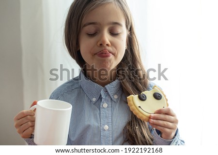 An elementary school girl is having breakfast with milk and funny cookies in the form of a smiley.