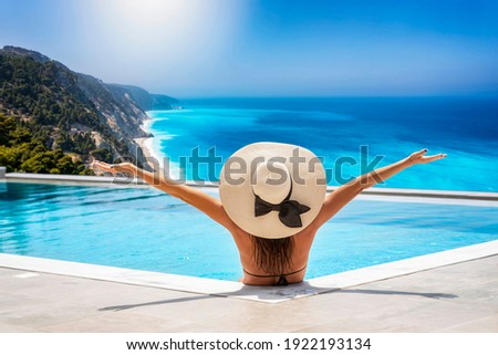 A happy woman in bikini and with hat sits in the swimming pool and enjoys the beautiful view to the turquoise sea of Lefkada island, Ionian Sea, Greece, during summer time Royalty-Free Stock Photo #1922193134