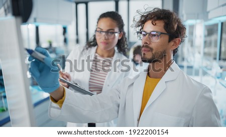 Medical Science Laboratory: Handsome Latin Male Scientist Writes Detailed Project Data Analysis on the Board, His Black Female Colleague Talks. Young Scientists Solving Problems. Royalty-Free Stock Photo #1922200154