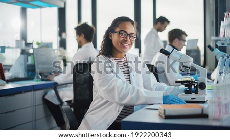 Medical Science Laboratory: Beautiful Black Scientist is Using Microscope, Looking at Camera and Smiling Charmingly. Young Biotechnology Science Specialist, Using Technologically Advanced Equipment. Royalty-Free Stock Photo #1922200343
