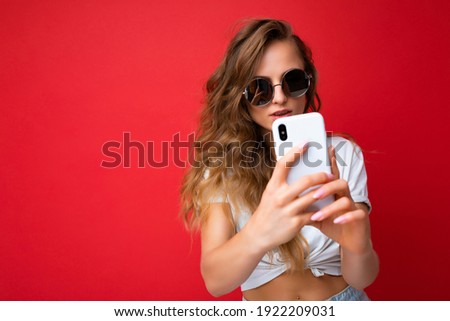 beautiful young woman holding mobile phone taking selfie photo using smartphone camera wearing everyday stylish outfit isolated over colorful wall background looking at device screen