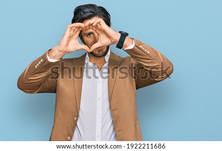 Young hispanic man wearing business clothes doing heart shape with hand and fingers smiling looking through sign 