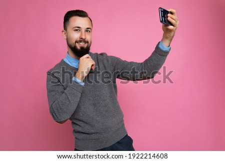 Photo of positive handsome young brunette unshaven man with beard wearing casual grey sweater and blue shirt isolated on pink background wall holding smartphone taking selfie photo looking at camera