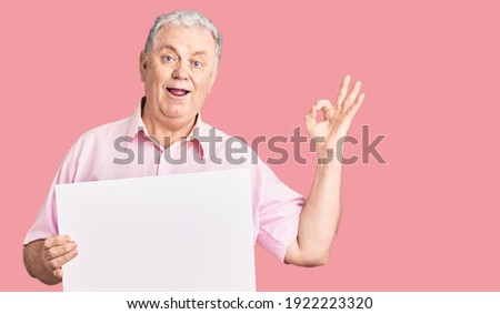 Senior grey-haired man holding blank empty banner doing ok sign with fingers, smiling friendly gesturing excellent symbol 