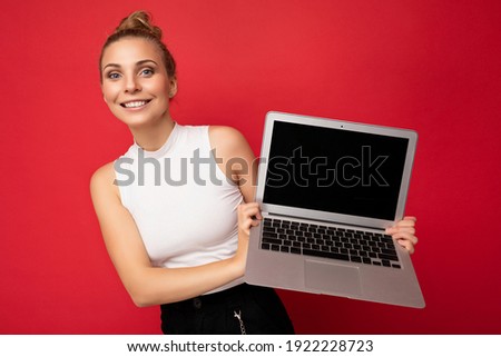 beautiful smiling fascinating happy blond young woman with gathered hair looking at camera holding computer laptop wearing white t-shirt isolated over red wall background