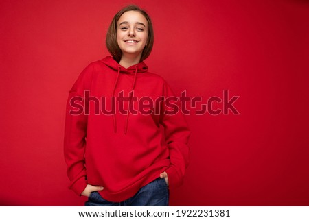 Photo shot of beautiful positive smiling brunette little girl wearing stylish red hoodie standing isolated over red background wall looking at camera