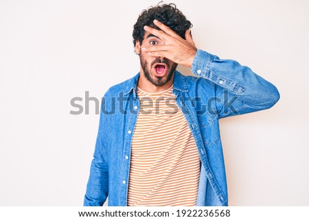 Handsome young man with curly hair and bear wearing casual denim jacket peeking in shock covering face and eyes with hand, looking through fingers with embarrassed expression.  Royalty-Free Stock Photo #1922236568