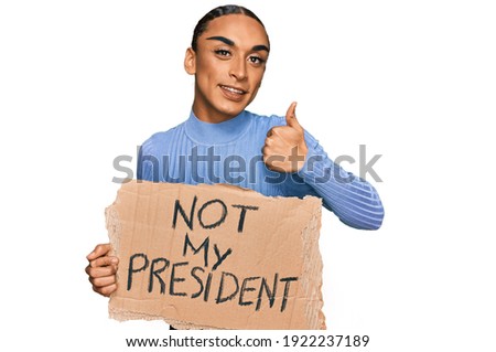 Hispanic transgender man wearing make up and long hair holding not my president protest banner smiling happy and positive, thumb up doing excellent and approval sign 