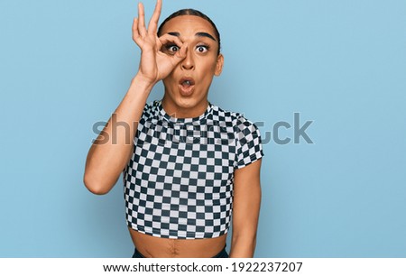 Hispanic transgender man wearing make up and long hair wearing modern clothes doing ok gesture shocked with surprised face, eye looking through fingers. unbelieving expression. 