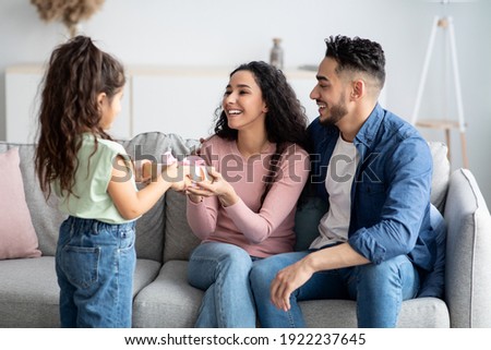 Mother's Day Greeting. Happy Young Arabic Woman Receiving Gift From Daughter And Husband At Home, Getting Pleasant Surprise From Her Child And Spouse, Sitting On Couch In Living Room, Free Space