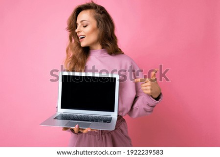 Photo of beautiful blond curly young feemale teenager girl lookind to the side holding computer laptop with empty monitor screen with mock up and copy space pointing at netbook with hand and one