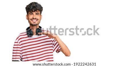 Young hispanic man listening to music using headphones gesturing with hands showing big and large size sign, measure symbol. smiling looking at the camera. measuring concept. 