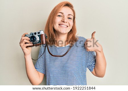 Young caucasian woman holding vintage camera smiling happy and positive, thumb up doing excellent and approval sign 