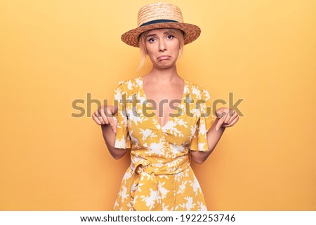 Beautiful blonde woman on vacation wearing summer hat and dress over yellow background Pointing down looking sad and upset, indicating direction with fingers, unhappy and depressed.