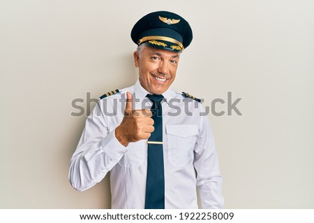 Handsome middle age mature man wearing airplane pilot uniform doing happy thumbs up gesture with hand. approving expression looking at the camera showing success. 