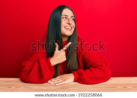 Beautiful hispanic woman wearing casual clothes sitting on the table cheerful with a smile of face pointing with hand and finger up to the side with happy and natural expression on face 