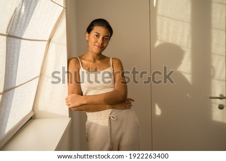 Waist up portrait of the dreamful girl relaxing at home while standing near the wall and smiling to the camera. Time at home concept. Stock photo