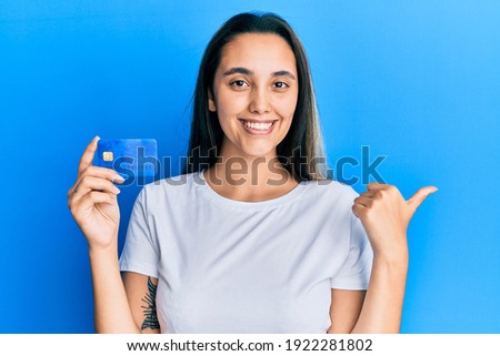 Young hispanic woman holding credit card pointing thumb up to the side smiling happy with open mouth 