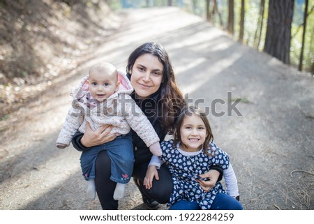 Portrait of happy woman holding her two adorable young daughters in the woods. Mother hugging a smiling little girl and cute baby on path with blurry trees as background. Young family in forest