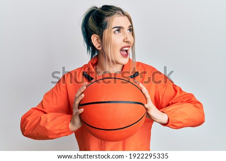 Young modern girl holding basketball ball angry and mad screaming frustrated and furious, shouting with anger looking up. 