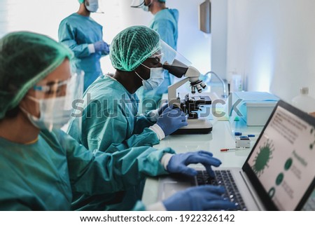 Multiracial medical scientists in hazmat suit working with microscope and laptop computer inside hospital lab - Focus african man face Royalty-Free Stock Photo #1922326142