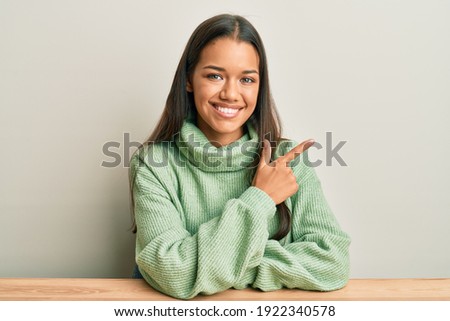 Beautiful hispanic woman wearing casual clothes sitting on the table smiling cheerful pointing with hand and finger up to the side 