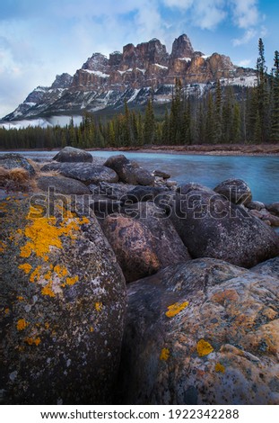 Orange lichen covered limestone rocks line the banks of the glacial blue water of the Bow River in Banff National Park, Alberta, Canada below the famous Castle Mountain.