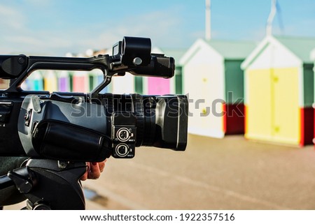 In focus and foreground a professional video camera
Out of focus colorful huts on the seafront at Brighton Beach, UK