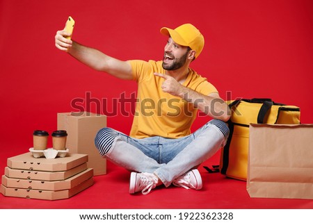 Delivery employee man guy male in yellow cap t-shirt sit at thermal takeaway food bag backpack pizza boxes cups hold mobile cell phone work courier service during covid-19 isolated on red background