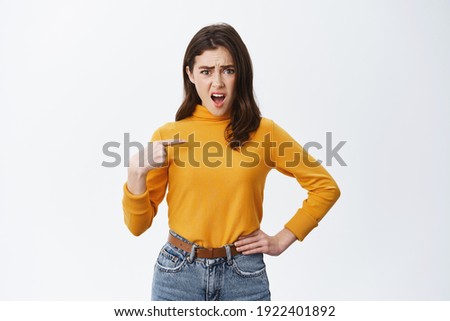 Insulted shocked young woman pointing at herself and gasping, being offended, staring at camera with displeased face, standing against white background.