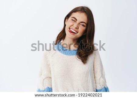 Positive emotions. Portrait of beautiful young brunette woman in sweater, winking and showing tongue while smiling from joy and happiness, standing against white background.