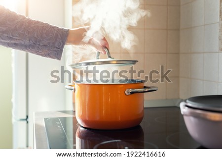 Female hand open lid of enamel steel cooking pan on electric hob with boiling water or soup and scenic vapor steam backlit by warm sunlight at kitchen. Kitchenware utensil and tool at home background Royalty-Free Stock Photo #1922416616