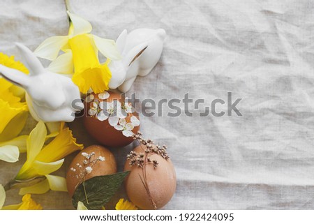 Happy Easter! Stylish Easter eggs decorated with dry flowers and petals on rustic linen napkin with yellow daffodils and white bunnies. Flat lay, Space for text. Creative natural eco friendly decor