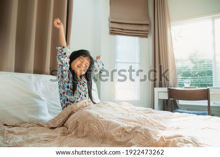 Asian child girl enjoys sunny morning. Good time at home. Kid wakes up from sleep. child girl wakes up in morning in bed and stretches by window. morning awakening little child girl in bed. Royalty-Free Stock Photo #1922473262