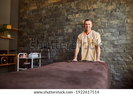 Copy-space photo of easy-going masseur standing near spa bed and looking at the camera