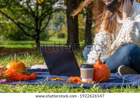 Selective focus. View of woman in white sweater and protective face mask sitting with laptop in autumn public park on blue rug. Orange pumpkins and coffee paper cup lies nearby. Outdoor work theme.
