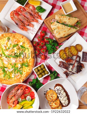 Traditional Italian cuisine concept flat lay. Sea food pizza, cooked shrimps, caramel ribs, ham pita sandwich, green olives and fresh cherry tomatoes over red plaid tablecloth.