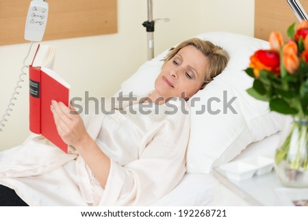 Attractive middle-aged woman lying in a hospital bed propped up on the pillows with a bowl of roses alongside her reading a book Royalty-Free Stock Photo #192268721