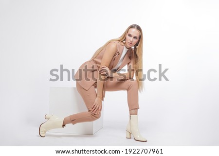 High fashion photo of a beautiful elegant young woman in a pretty beige suit, pants, vest, trousers, boots posing over white, soft gray background. Studio Shot, portrait. Model sits on a cube. Blonde